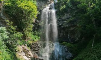 A beautiful waterfall surrounded by lush greenery and tall trees. The clear water flows into a small creek.