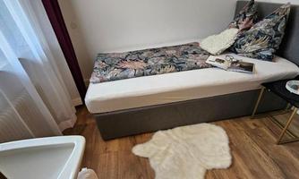 A cozy room with a single bed, decorated with colorful cushions. Next to the bed is a small table with a cup and a book.