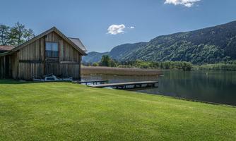 A wooden house by the shore of a tranquil lake, surrounded by green meadows and hills. The clear blue sky and the gentle landscape create a peaceful atmosphere.