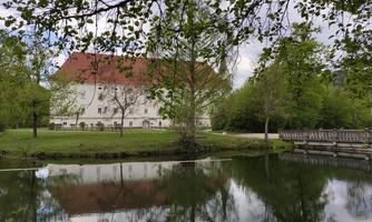 A beautiful, historic building with a red roof, surrounded by trees and a tranquil pond. The green landscape is reflected in the water.