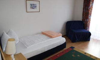 A simple bedroom with a single bed, a blue armchair, and a rug. The room has large windows with curtains and a cozy atmosphere.