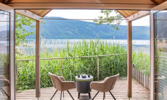 A cozy area with two chairs and a table that offers a view of a tranquil lake. Surrounded by lush greenery and windows that make the space bright and inviting.