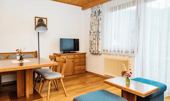 A bright room with wood decor, a dining table, and two cozy chairs. A television and a flower decoration give the room an inviting atmosphere.