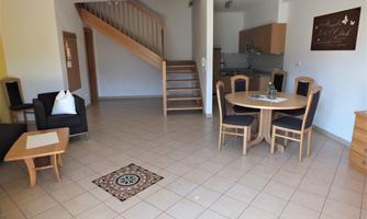 A bright and inviting living room with a staircase and a dining area. The furniture is simple and modern, featuring a small table and chairs.