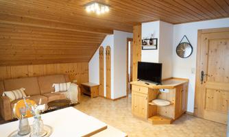 A cozy living space with wooden walls and a sloped ceiling. The room features a sofa, a television, and a dining table.