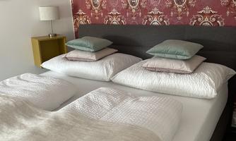 A cozy bedroom with a large bed and several pillows. The wall is decorated with an elegant patterned wallpaper design.