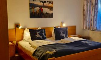 A cozy bedroom with a double bed and stylish cushions. A picture of a landscape hangs on the wall, and the window lets in soft light.