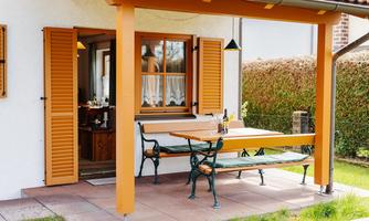 A cozy terrace with a wooden table and benches. The windows are decorated with orange shutters, surrounded by green plants.