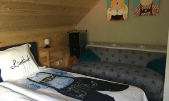 A cozy room with wooden paneling. On the bed lies a printed blanket and colorful pictures hang on the wall.