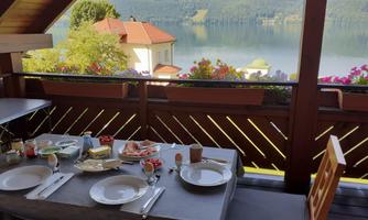 A beautiful breakfast table on a balcony overlooking a lake. Flowers and houses complement the picturesque view.