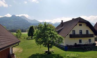 An idyllic landscape with a house, a large green garden, and mountains in the background. The sun is shining and the sky is partially cloudy.