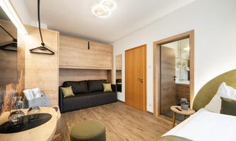 A modern hotel room with a comfortable bed, a seating area, and wooden shelves. The walls are in neutral colors and the floor is made of wood.
