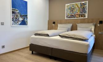 A modern bedroom with a double bed and white pillows. Two colorful artworks are hanging on the walls.