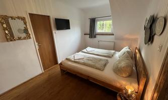 A cozy bedroom with a double bed and wooden floor. There is a window, a television, and decorative elements on the wall.