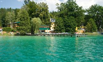 A picturesque lake landscape with clear, turquoise water and a waterfront promenade. Surrounded by lush greenery and some buildings by the water.