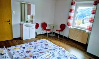 A simple room with a bed, a small table, and two red chairs. A window offers a view of the landscape.