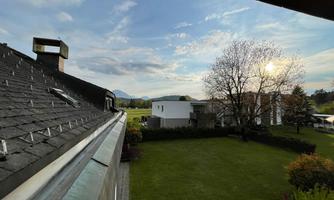 A view from a roof overlooking a meadow and a few houses. The sky is partly cloudy, and the sun is shining through the clouds.