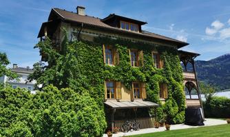 A beautiful house covered in green ivy. In the background, gentle hills and a clear blue sky can be seen.