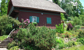 A rustic wooden house surrounded by colorful flowers and green plants. The garden is well-maintained and invites you to linger.
