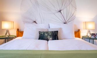 A cozy bedroom with a large double bed and white bedding. On the sides, there are nightstands with lamps and a decorative pillow.