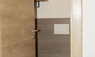 A modern bathroom with a white toilet and a wooden bathroom door. Shelves with toilet paper and cleaning supplies are mounted on the wall.