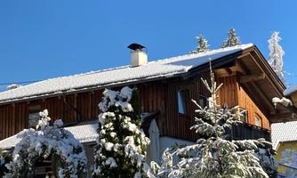 A beautiful wooden house with a snow-covered roof. The sky is clear and blue, surrounded by snow-covered trees.