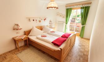 A cozy bedroom with a wooden bed and red pillows. Large windows with green curtains provide plenty of daylight.