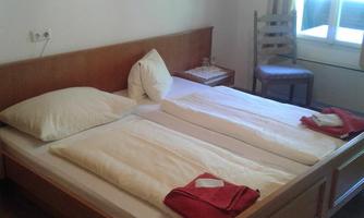 A simple bedroom with a double bed and two pillows. A window lets in a lot of light, and a chair stands next to the bed.
