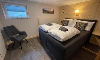 A cozy bedroom with a double bed and modern pillows. Next to the bed stands a comfortable armchair and there is warm lighting in the surroundings.