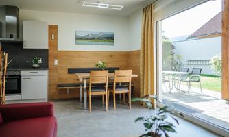 A modern kitchen with a dining area of wooden chairs. Large windows offer a view of a garden.