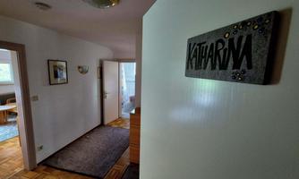 A bright hallway with a sign that says "KATHARINA." Pictures hang on the walls, and a carpet lies on the floor.