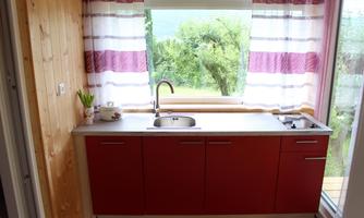A modern kitchen with red furniture and a large window. The windows are decorated with striped curtains and offer a view of nature.