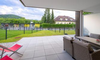 An inviting outdoor area with modern furniture and a beautiful view of the garden. In the background, trees and buildings can be seen.
