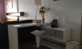 A modern living area with a white desk and an elegant chair. On the table, there is a coffee machine and a vase with flowers.