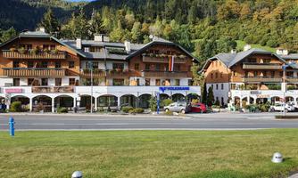 A charming hotel building with traditional architecture, surrounded by green mountains. In the foreground, cars and a well-kept lawn can be seen.
