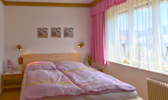 A cozy bedroom with a double bed and pink bedding. Large windows let in plenty of light and create a bright atmosphere.