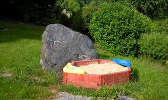 A sandbox with blue and yellow play equipment on a green lawn. In the background, there is a large rock and abundant vegetation.