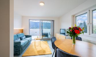 A modern living room with a blue couch, a table, and a beautiful view of the water. Bright colors and windows provide plenty of daylight.