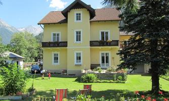 A beautiful yellow house with several balconies surrounded by a well-maintained garden. In the background, trees and mountains can be seen.