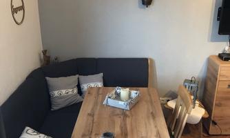 A cozy dining area with a wooden table and a seating area. The decor is modern and inviting with cushions and decorative elements.