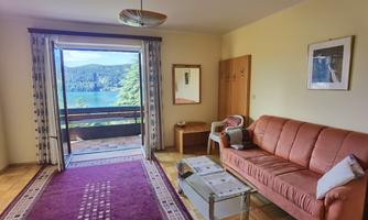 A cozy living room with a red sofa and a table. From the balcony, there is a beautiful view of the nature and the lake.