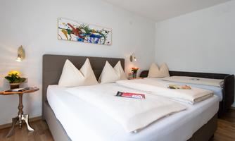A modern bedroom with two beds and decorative pillows. A colorful artwork hangs on the wall, and a small table stands next to a plant arrangement.