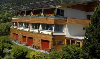 A modern building with a wooden façade and large windows, surrounded by green trees. The surroundings are hilly and provide a tranquil atmosphere.