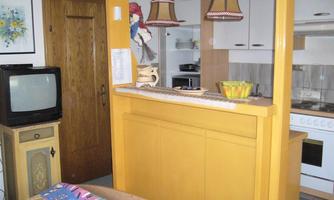 A cozy living kitchen with a yellow bar, a modern kitchen, and a television. The walls are decorated with artworks and there is a colorful bedspread in the foreground.