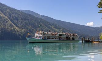 A boat is sailing on a clear, blue lake, surrounded by green mountains. Many people are enjoying the view on the boat.