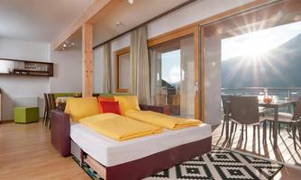 A bright and modern living room with a cozy sleeping area. Large windows offer a view of a sunny terrace with mountain views.