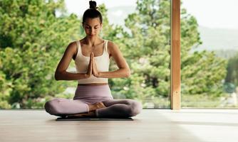 A woman is sitting in a lotus position and meditating in a bright room with large windows. Behind her, green trees are visible, creating a serene atmosphere.
