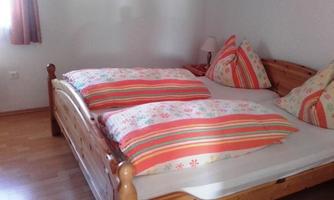 A cozy bedroom with a wooden bed and colorful cushions. The room has natural light and a warm wooden floor.