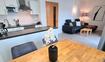 A modern apartment with an open kitchen and a cozy living area. The dining table is made of wood and a blooming plant is in the center.