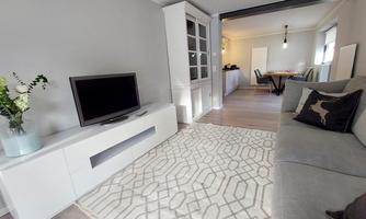 A modern living room with a gray sofa and a television. The floor is decorated with a light rug, and there is a dining area in the background.